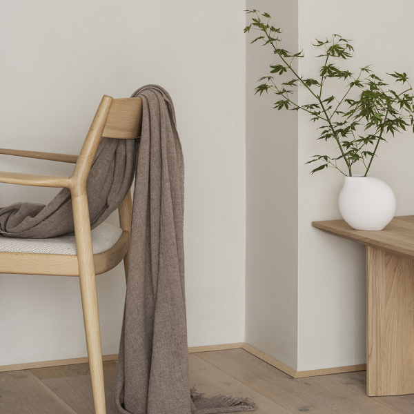 Cozy interior with a chair, draped blanket, and a vase of greenery on a wooden table.