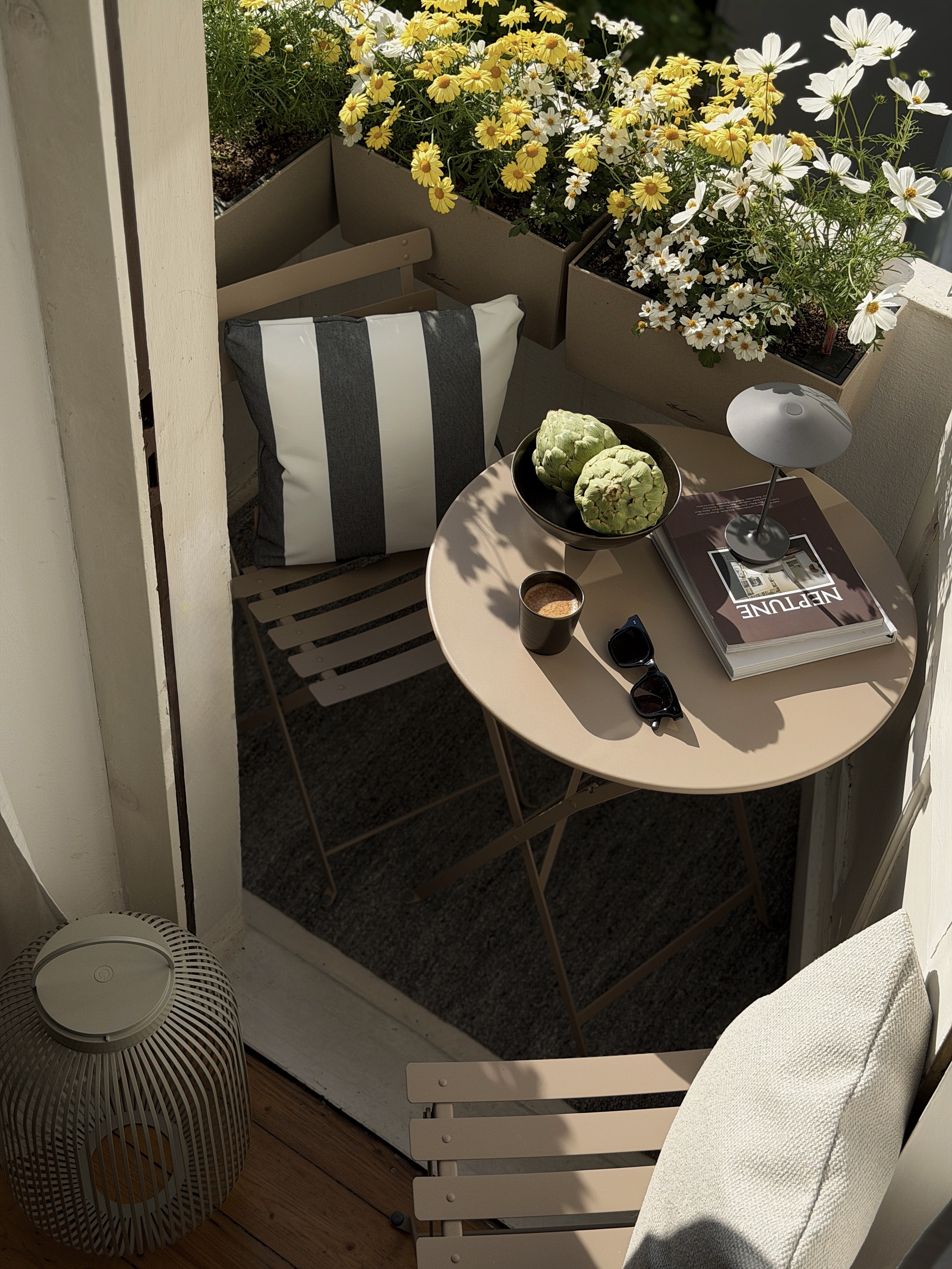 Kleiner sonniger Balkon mit beigefarbenem Tisch, Stühlen, gestreiftem Kissen, Blumenkästen und dekorativen Outdoor-Accessoires.