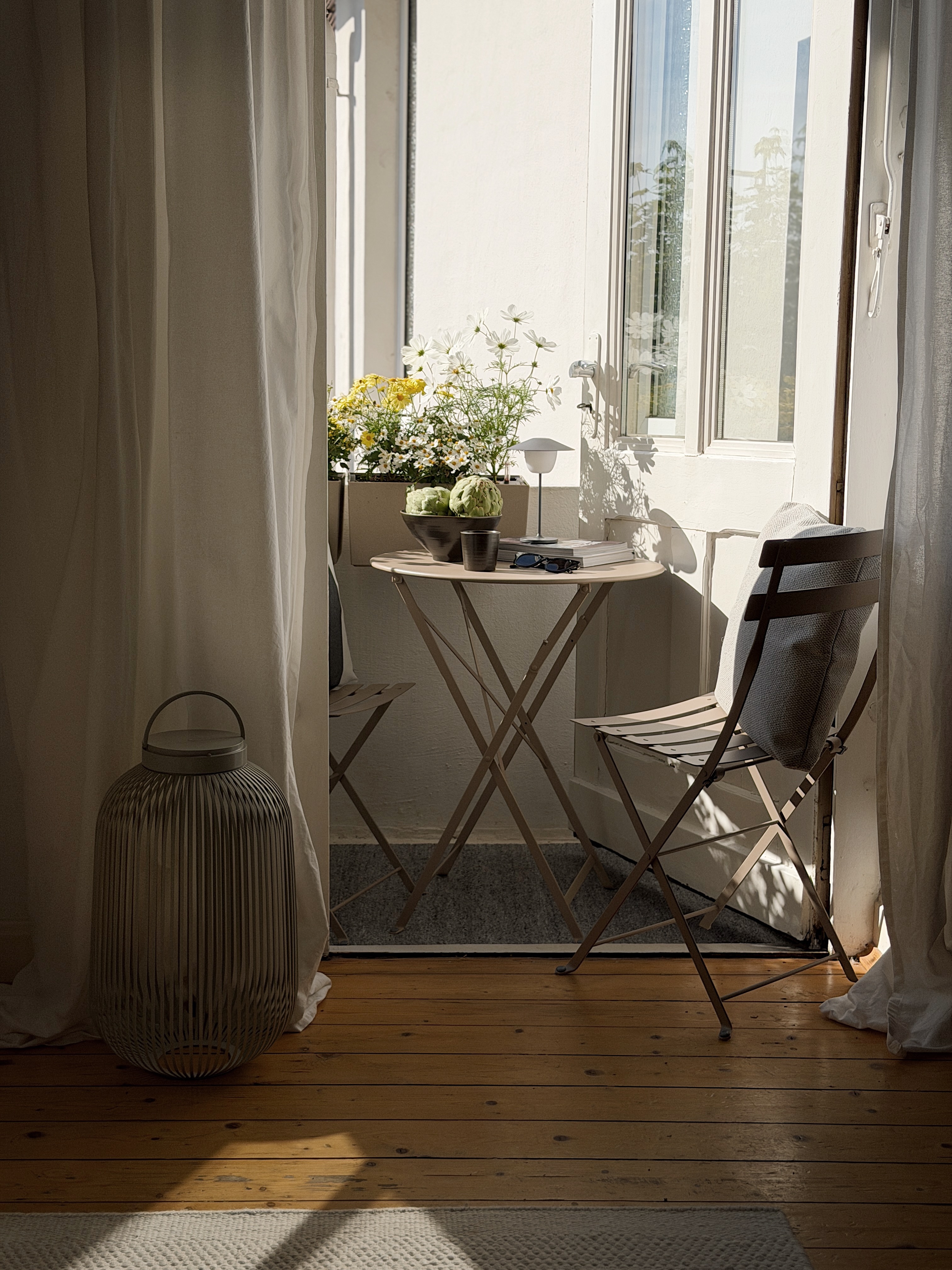 Gemütlicher kleiner Balkon mit beigem Klapptisch, Stühlen, Blumenkästen, dekorativer Laterne und sanftem Sonnenlicht.