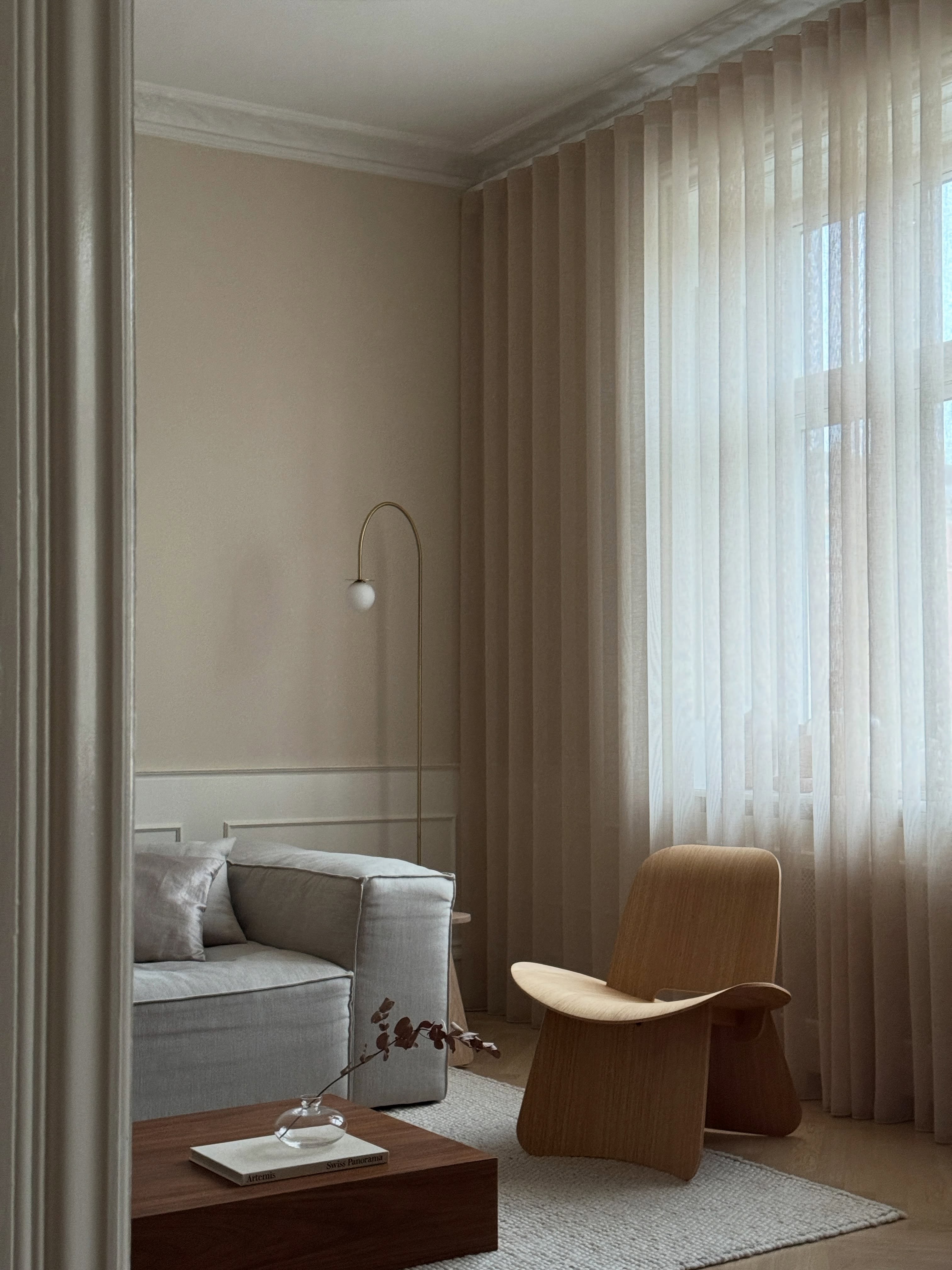 Wohnzimmer im Japandi-Stil mit grauem Sofa, geschwungenem Holzsessel, Holzcouchtisch und bodenlangen beigen Vorhängen.