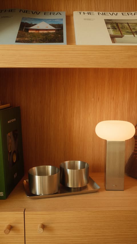 Warm leuchtende Tischlampe neben zwei Metallgefäßen auf einem Tablett in einem Holzregal mit Büchern.