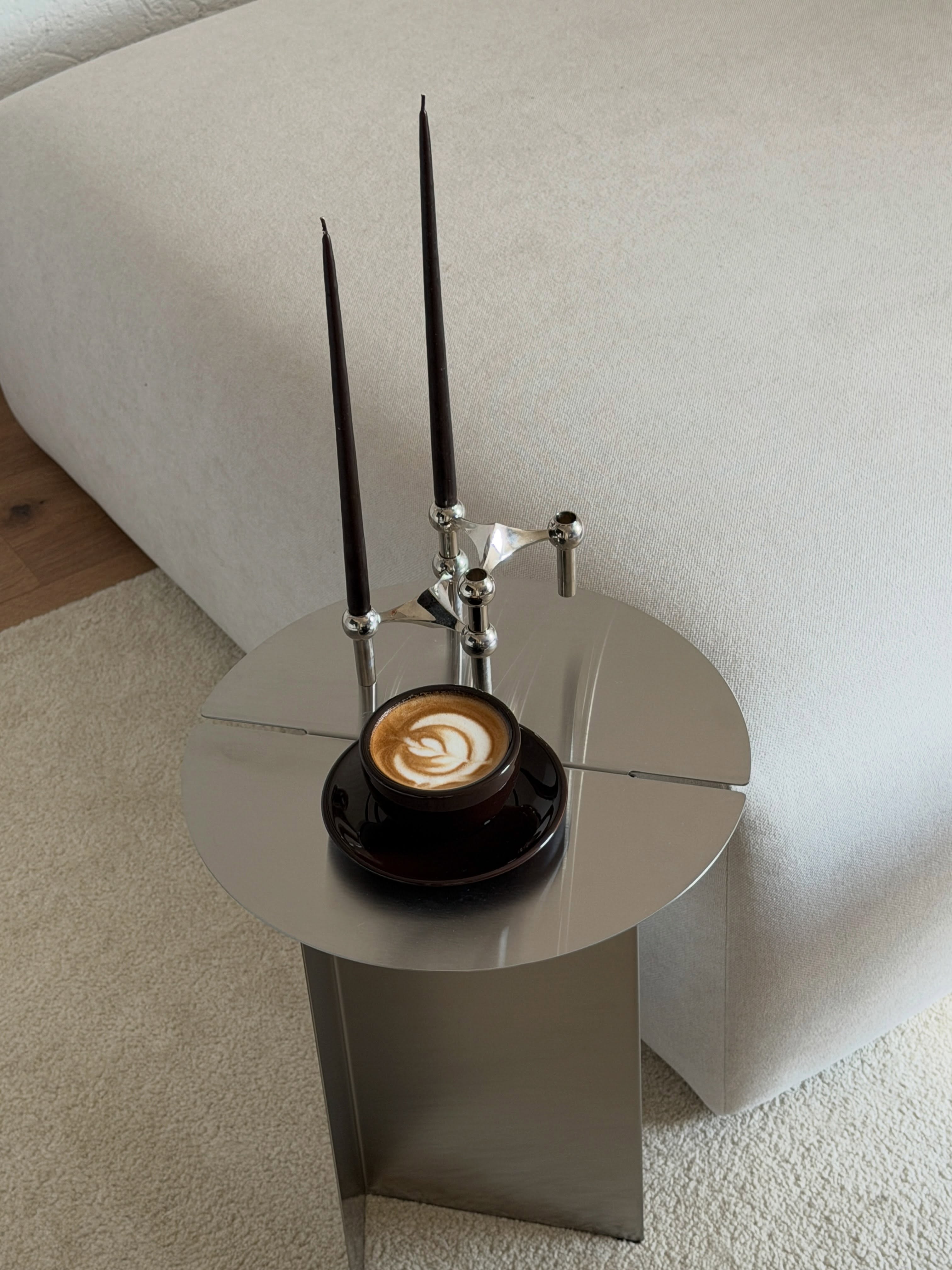 Runder Metall-Beistelltisch mit Cappuccino und zwei schwarzen Stabkerzen in silbernen Kerzenhaltern neben einem hellen Sofa.