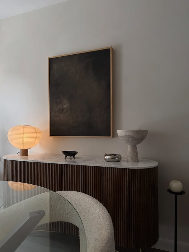 Dunkles Holzsideboard mit heller Steinplatte, leuchtender Papierlampe und dekorativen Schalen unter einem abstrakten Wandbild.