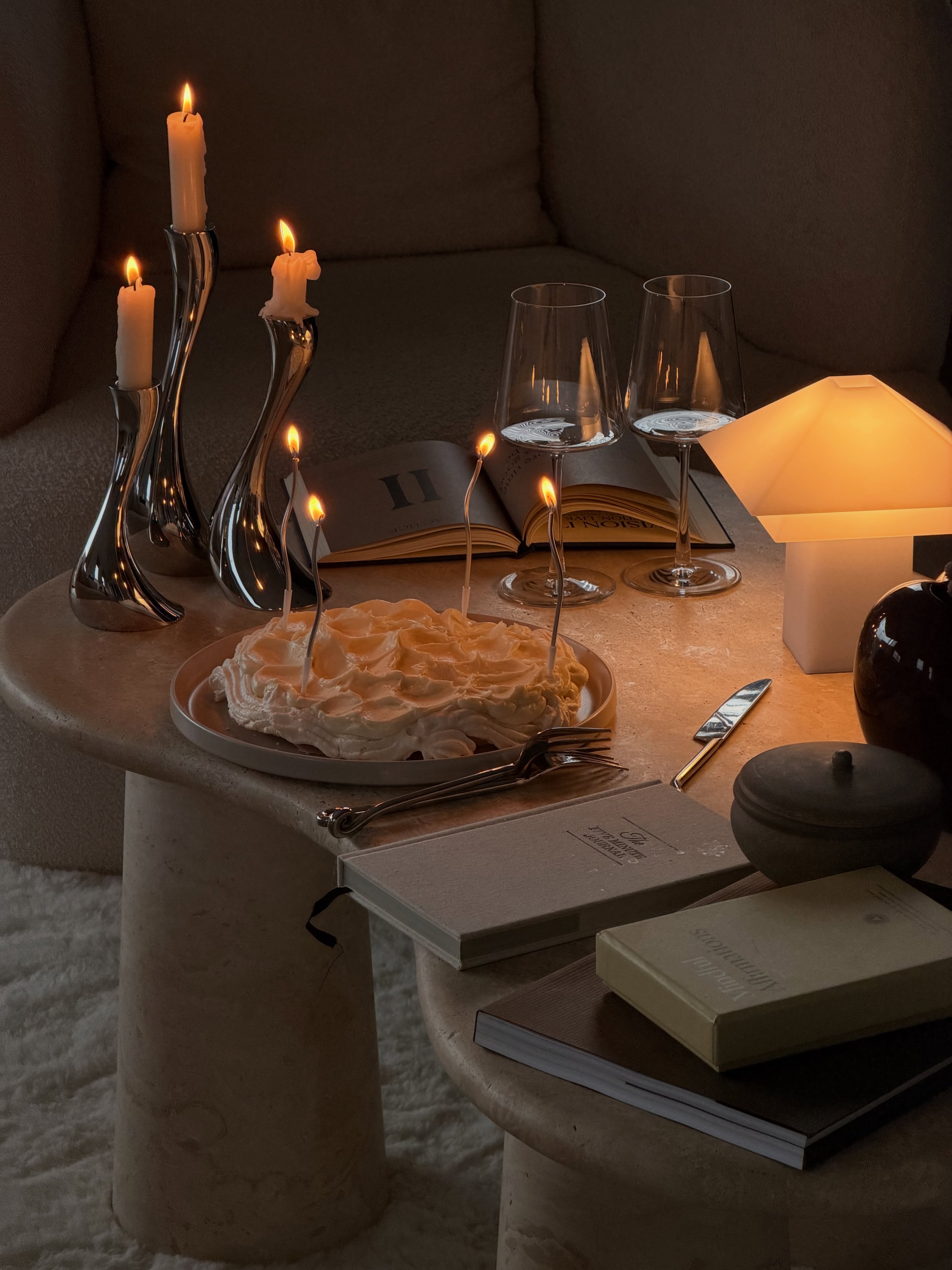 Bougeoirs argentés, lampe de table allumée et gâteau avec des bougies allumées sur une table basse beige.